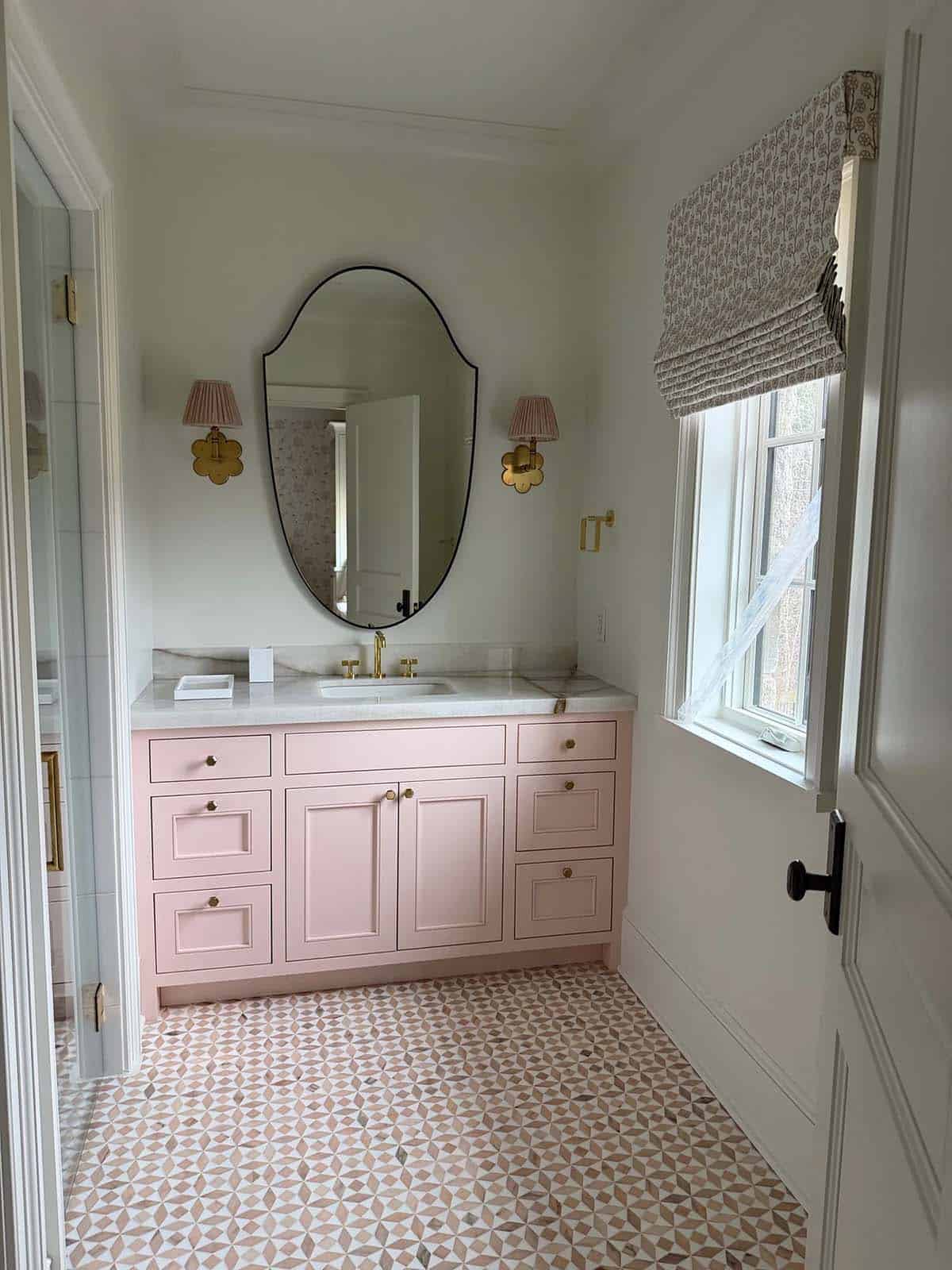 Pink bathroom vanity with oval mirror, brass sconces, and patterned tile floor