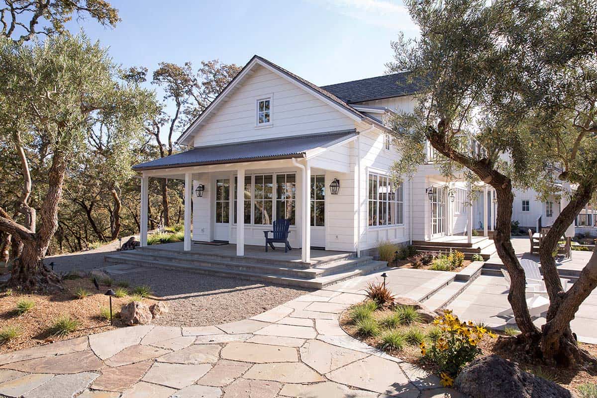 White farmhouse guest wing with covered porch, gravel path, and olive trees