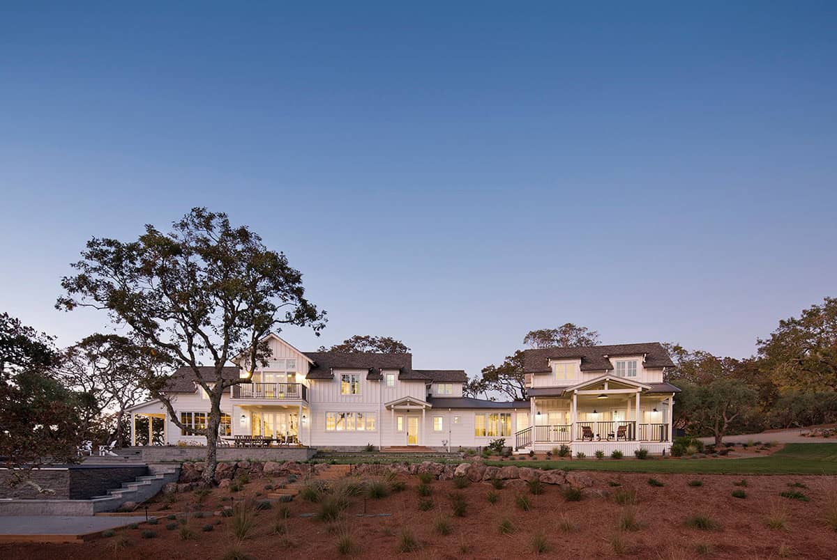 Twilight view of the illuminated farmhouse with covered porches and stacked-stone patio