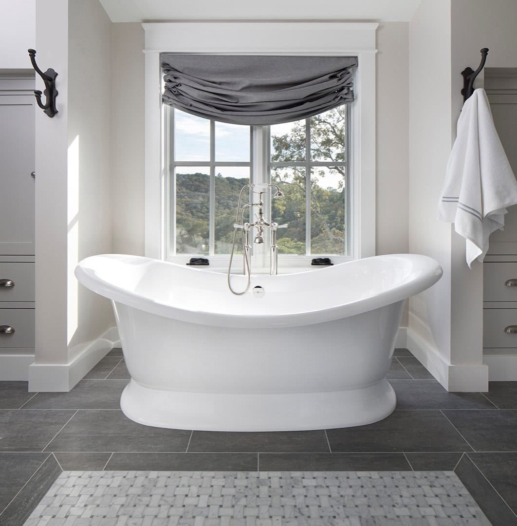 Freestanding soaking tub beneath a window with gray Roman shade and hillside view