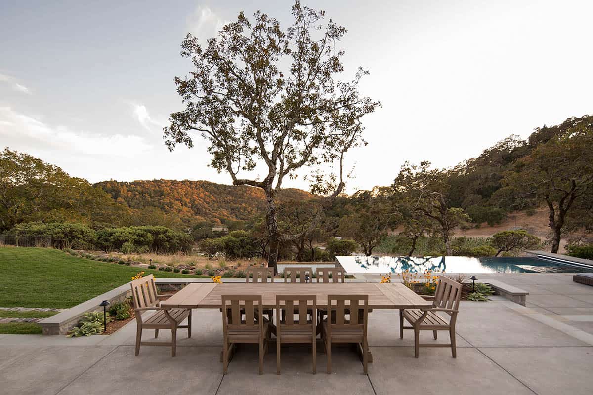 Outdoor dining table on a patio overlooking an infinity pool and wooded hills at dusk