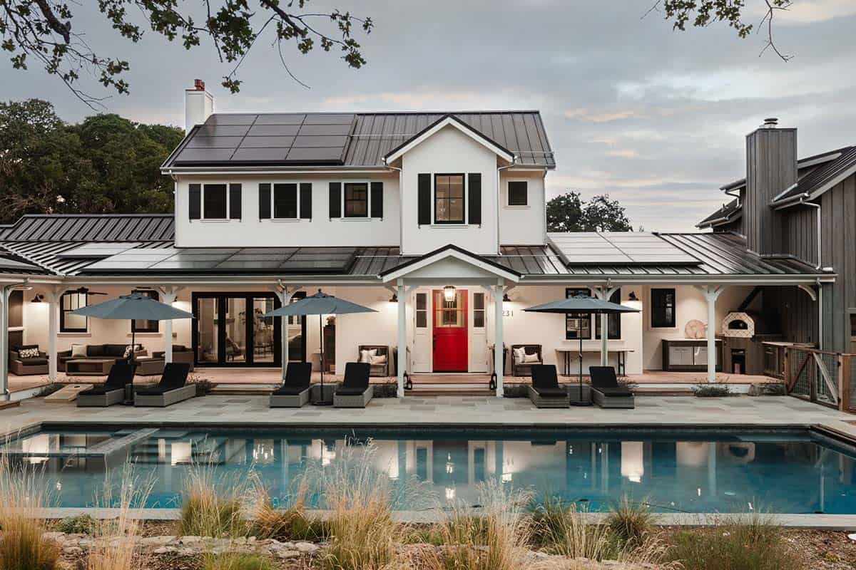 White modern farmhouse exterior with black metal roof, red front door, and pool at dusk