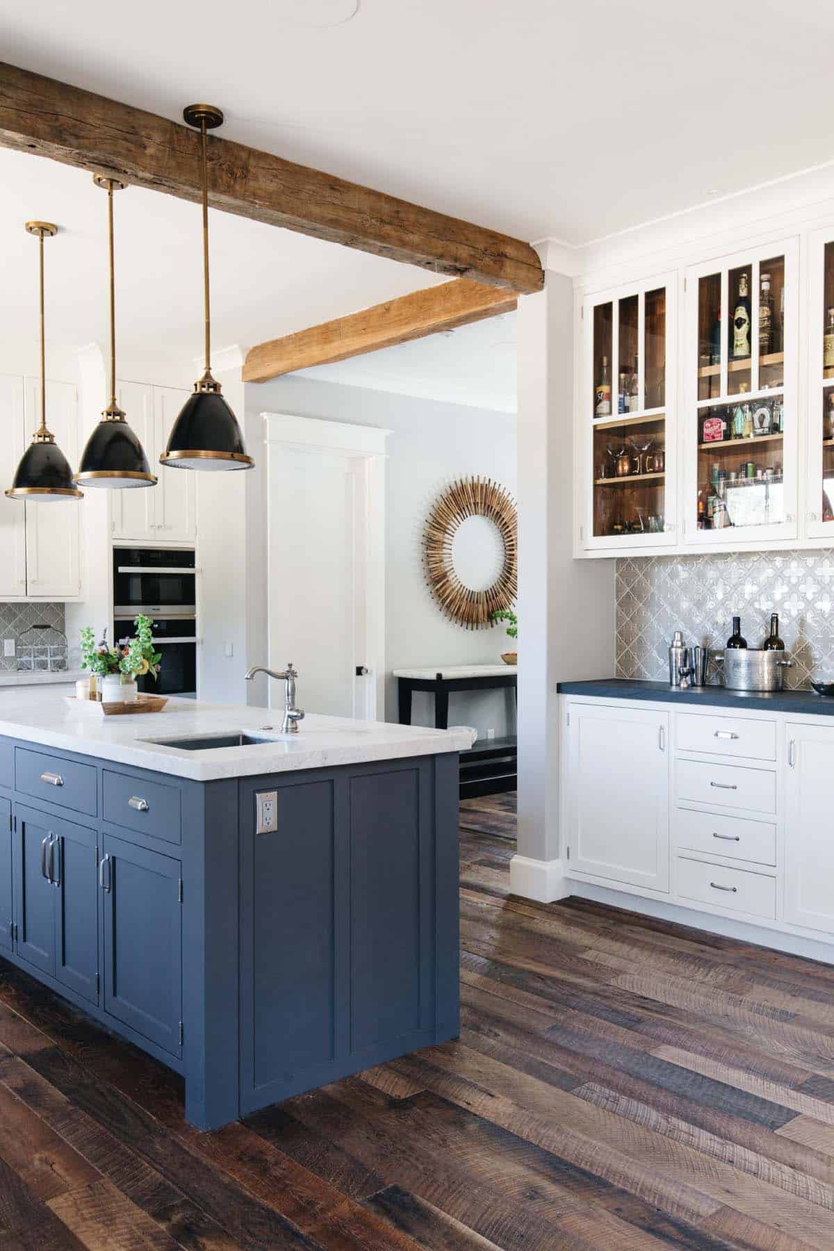 Kitchen island with navy cabinetry next to a glass-front bar cabinet and reclaimed wood beam