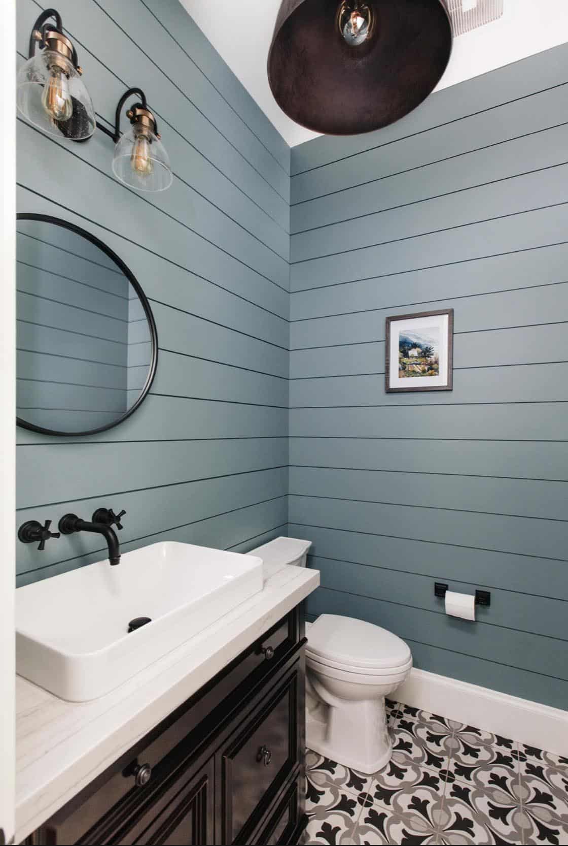 Powder room with blue shiplap walls, round black mirror, and patterned tile floor