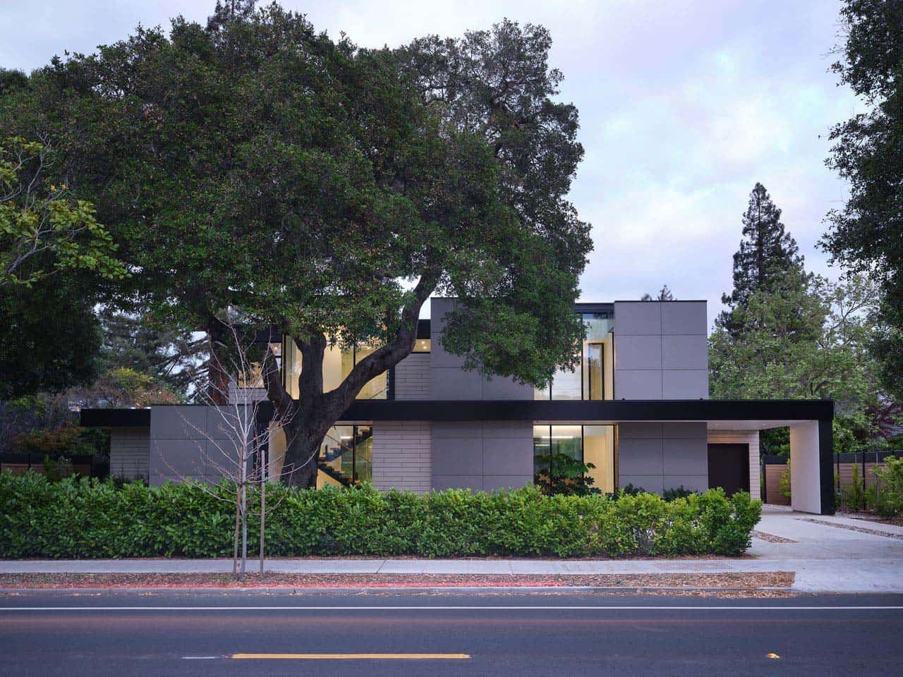 Evening street view of the home with illuminated interior behind the oak tree