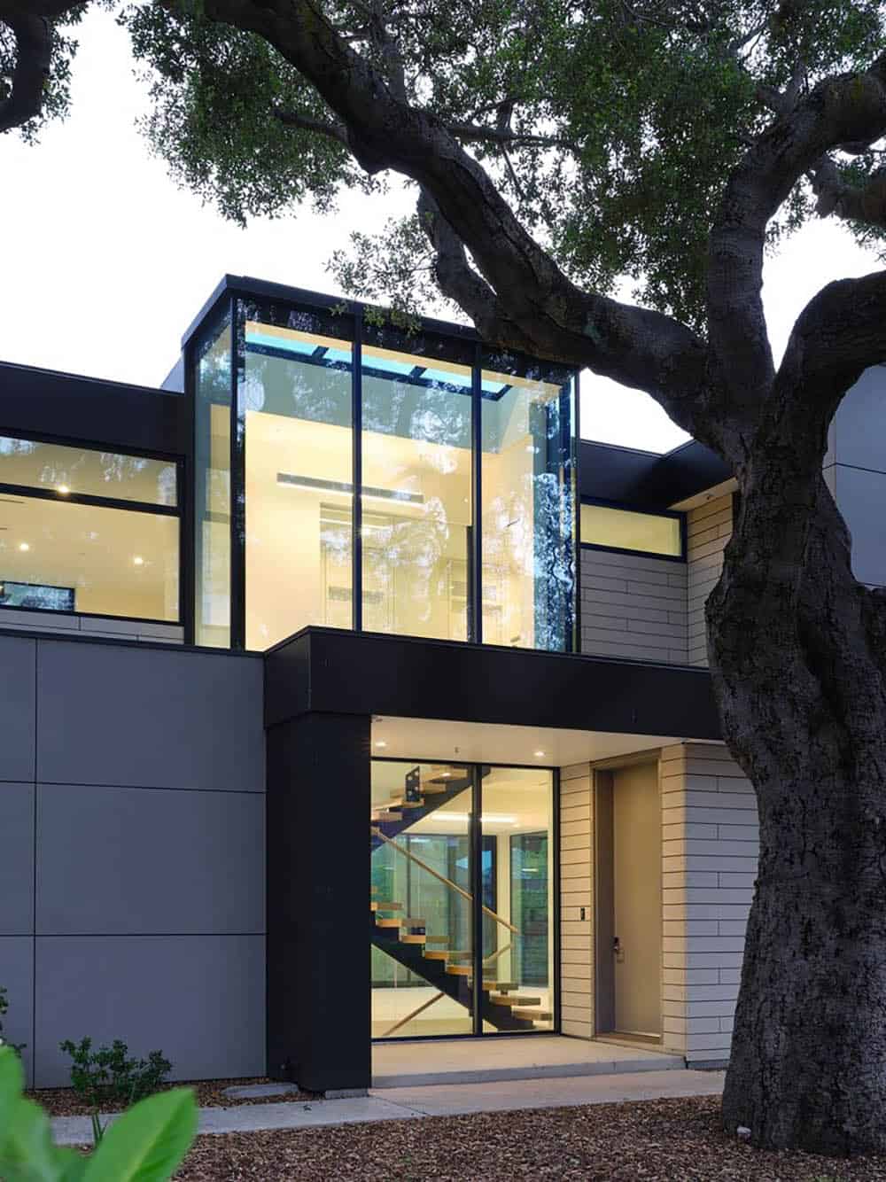 Twilight view of the glass stairwell lantern and entry beneath the oak tree