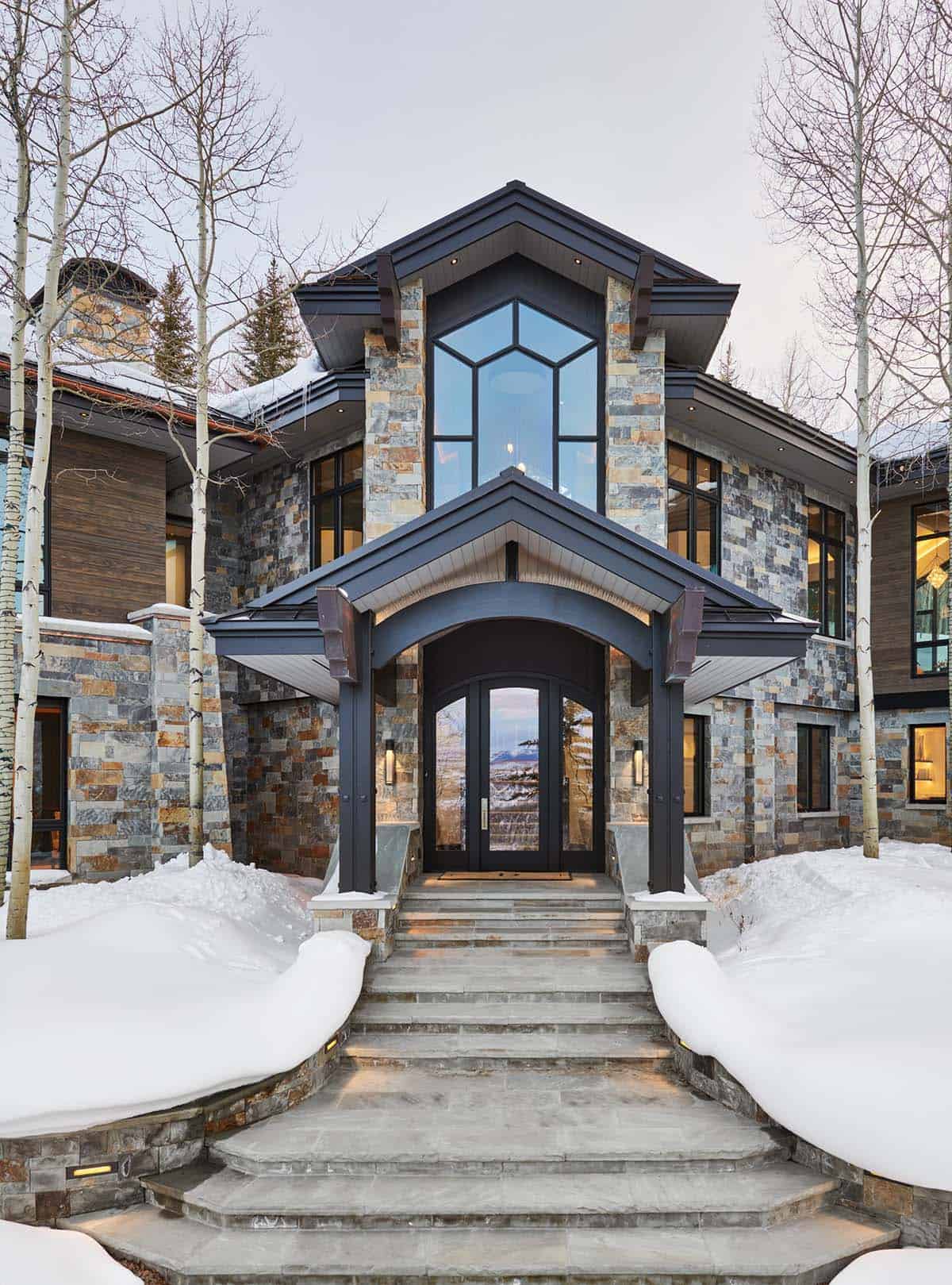 Home entrance with stone columns, arched dark door, and snow-lined stairway