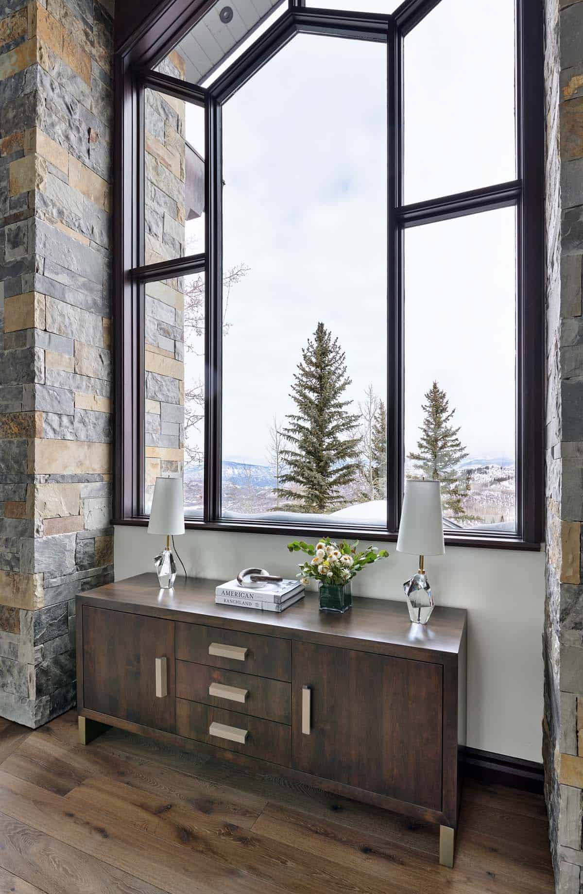 Wood console table beneath tall angular windows with snowy pine view