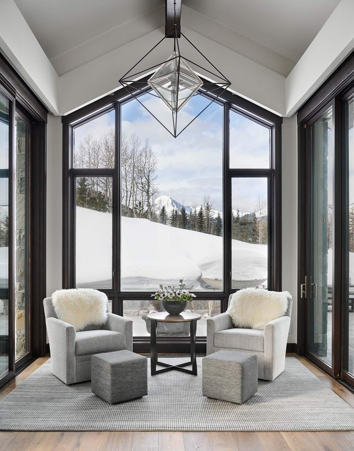 Sunroom sitting area with swivel chairs, ottomans, and geometric pendant light overlooking snow.