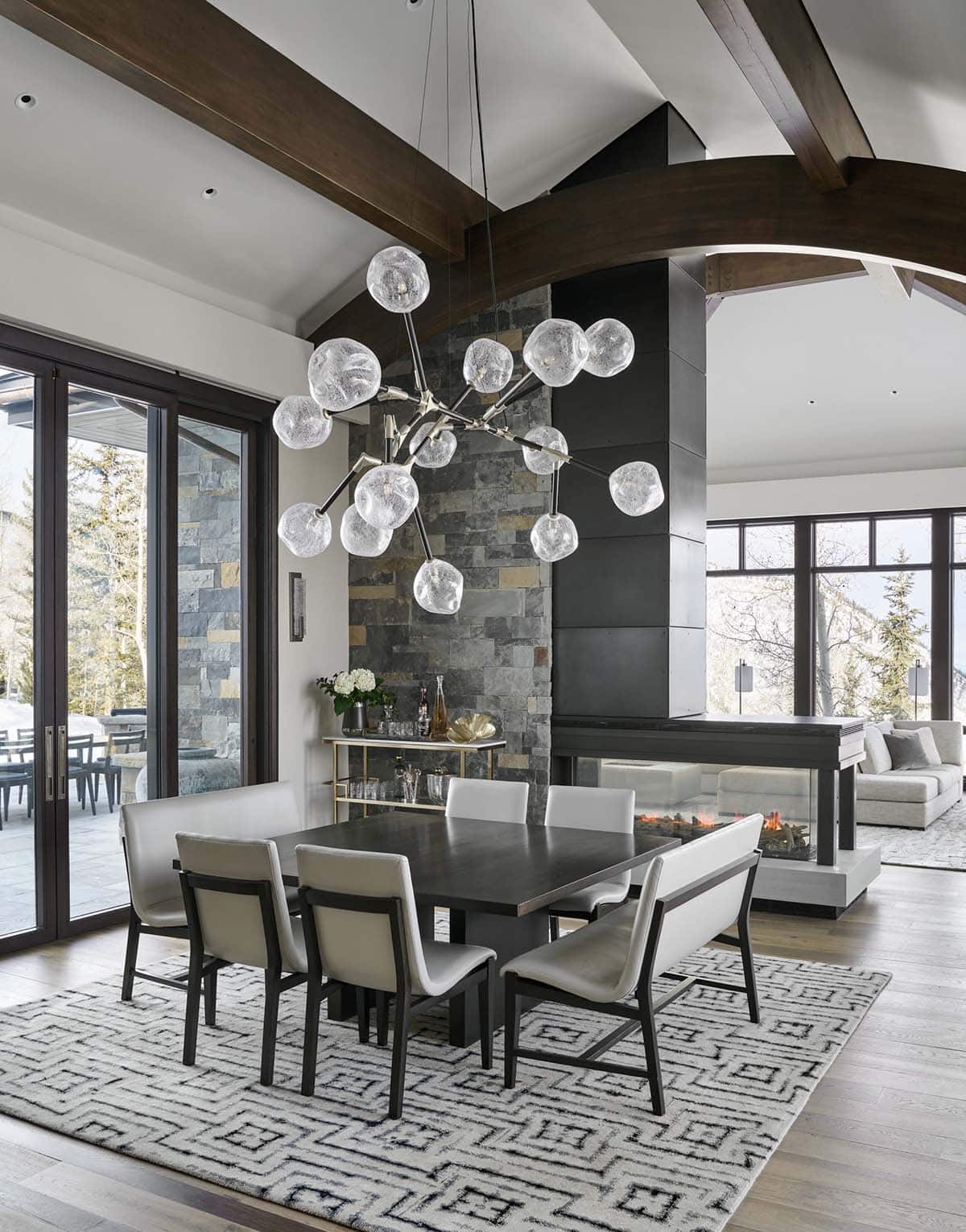 Dining area with sculptural glass-orb chandelier and double-sided stone fireplace