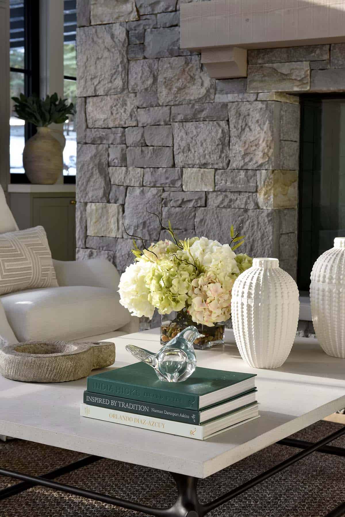 Coffee table styled with books, glass bird figurine, and white vases near stone fireplace