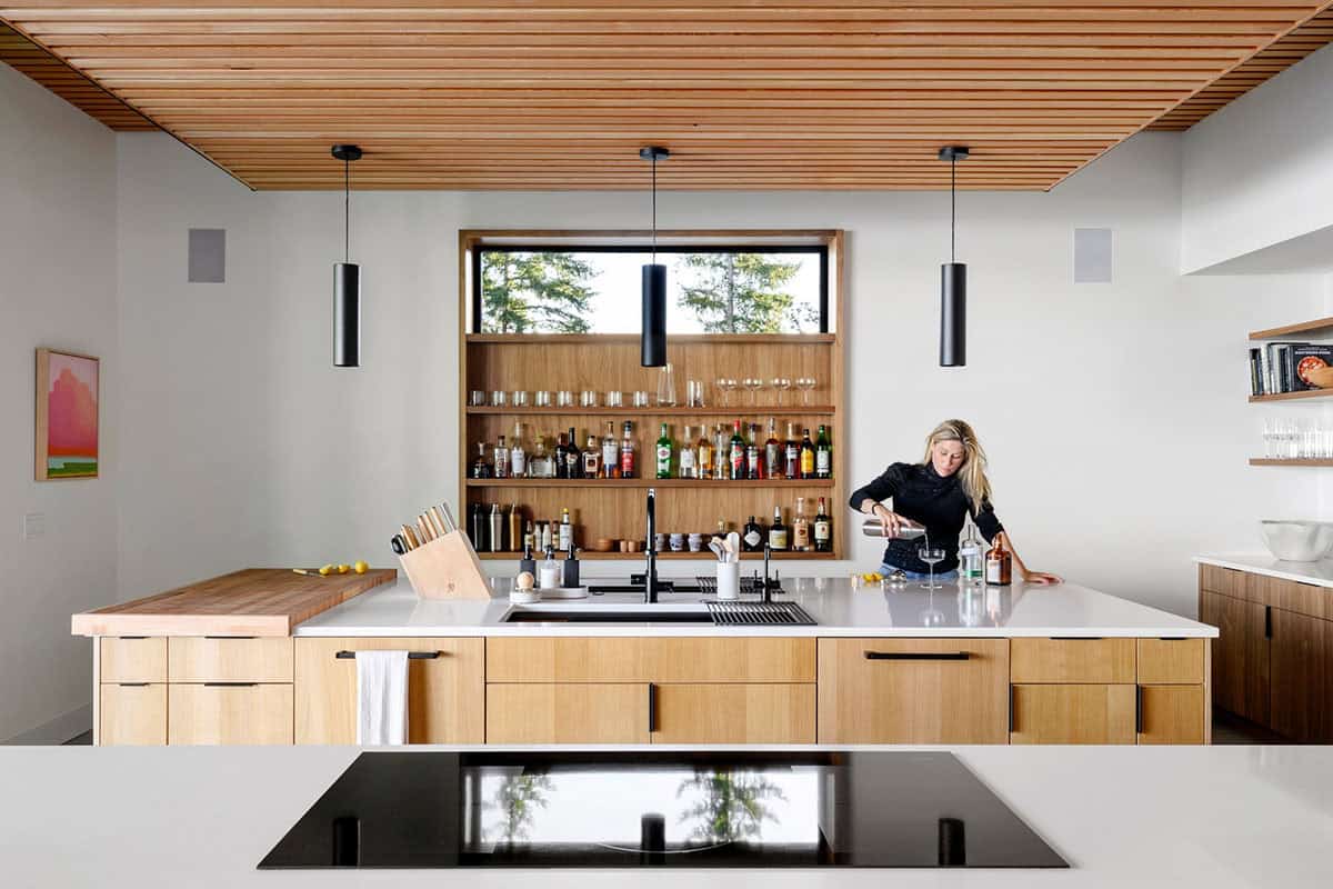 Wife pours cocktails at wood island beneath slatted ceiling and bar nook