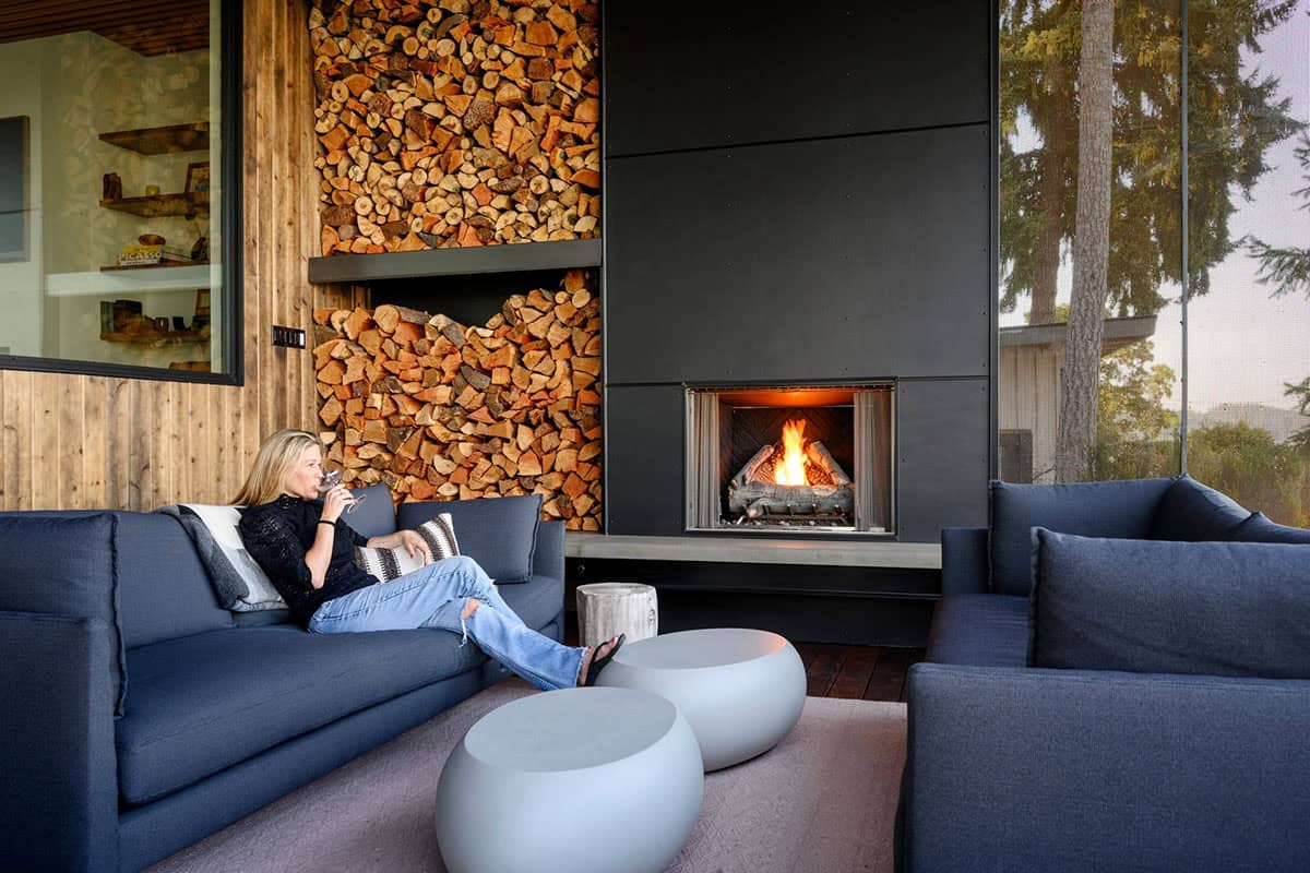 Woman relaxes by fireplace beside stacked firewood storage wall