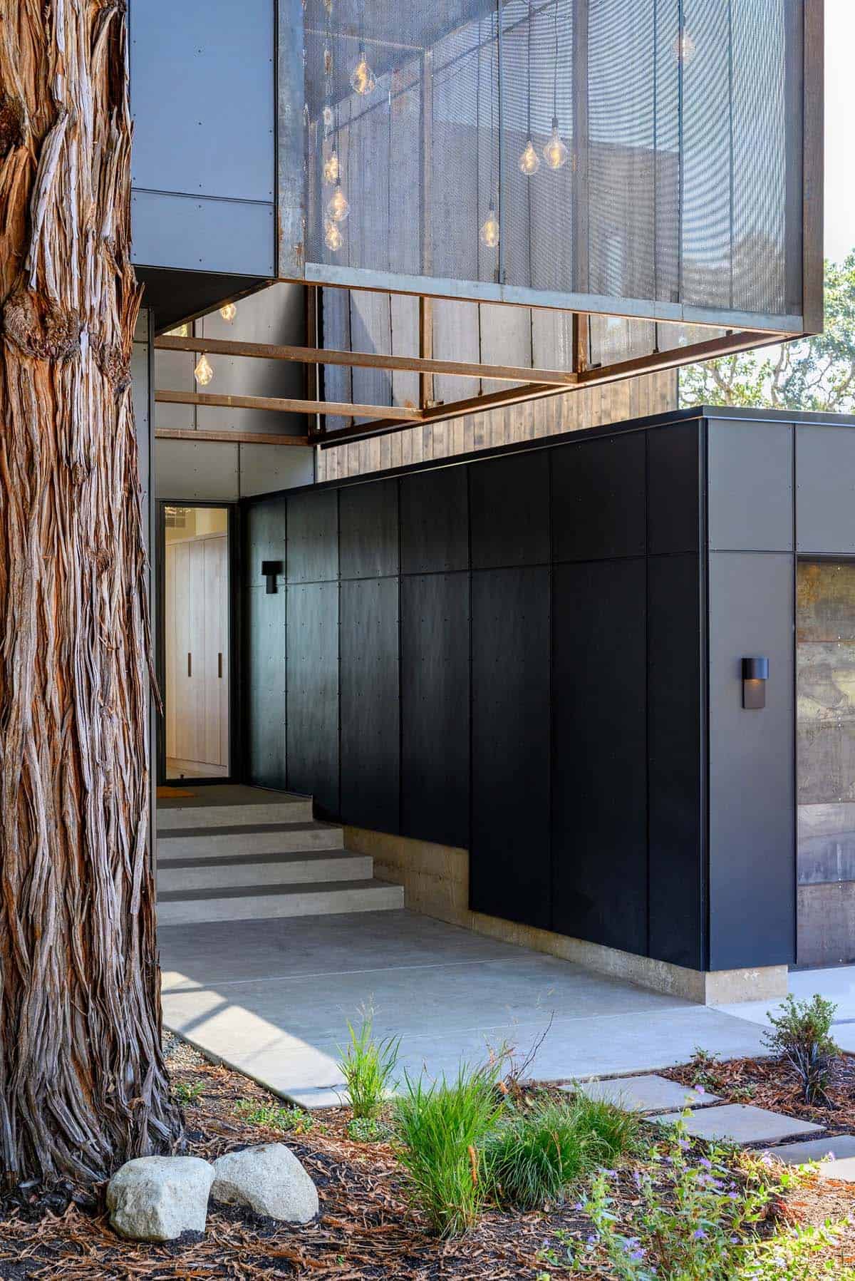 Black paneled entry facade beside cedar tree trunk and steps