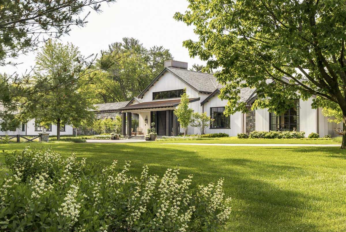 Front exterior of white board-and-batten farmhouse with cedar shake roof, framed by mature trees and lawn