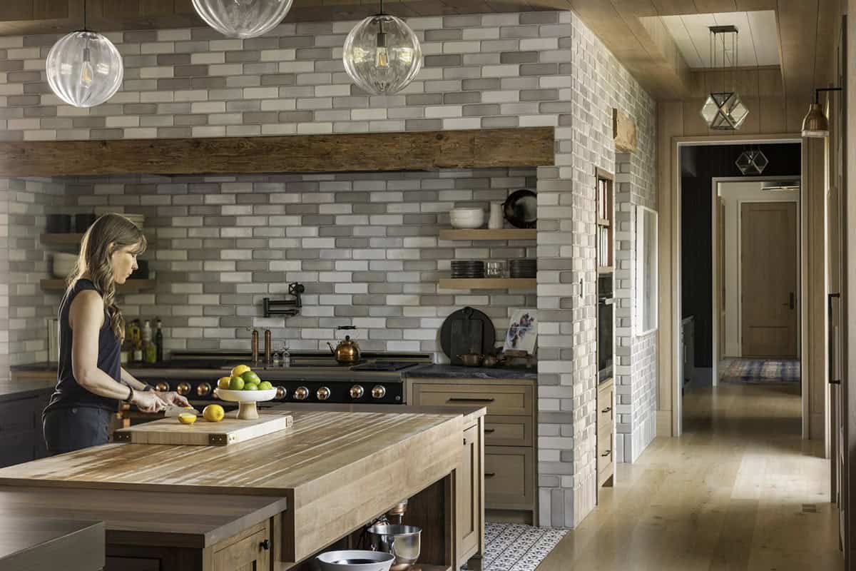 Woman preparing food at butcher block island beneath a brick-tiled range alcove with open shelving