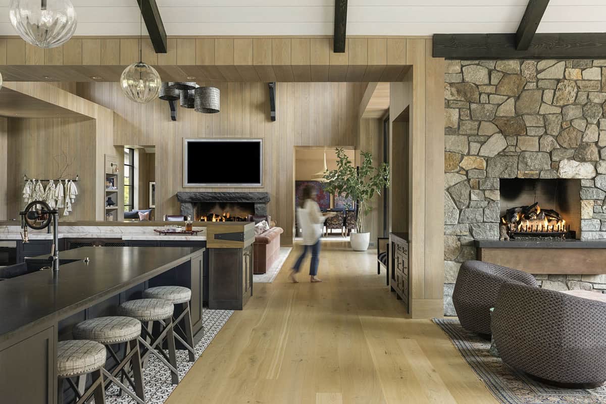View through kitchen toward stone fireplace, mounted TV, and figure walking down the hallway