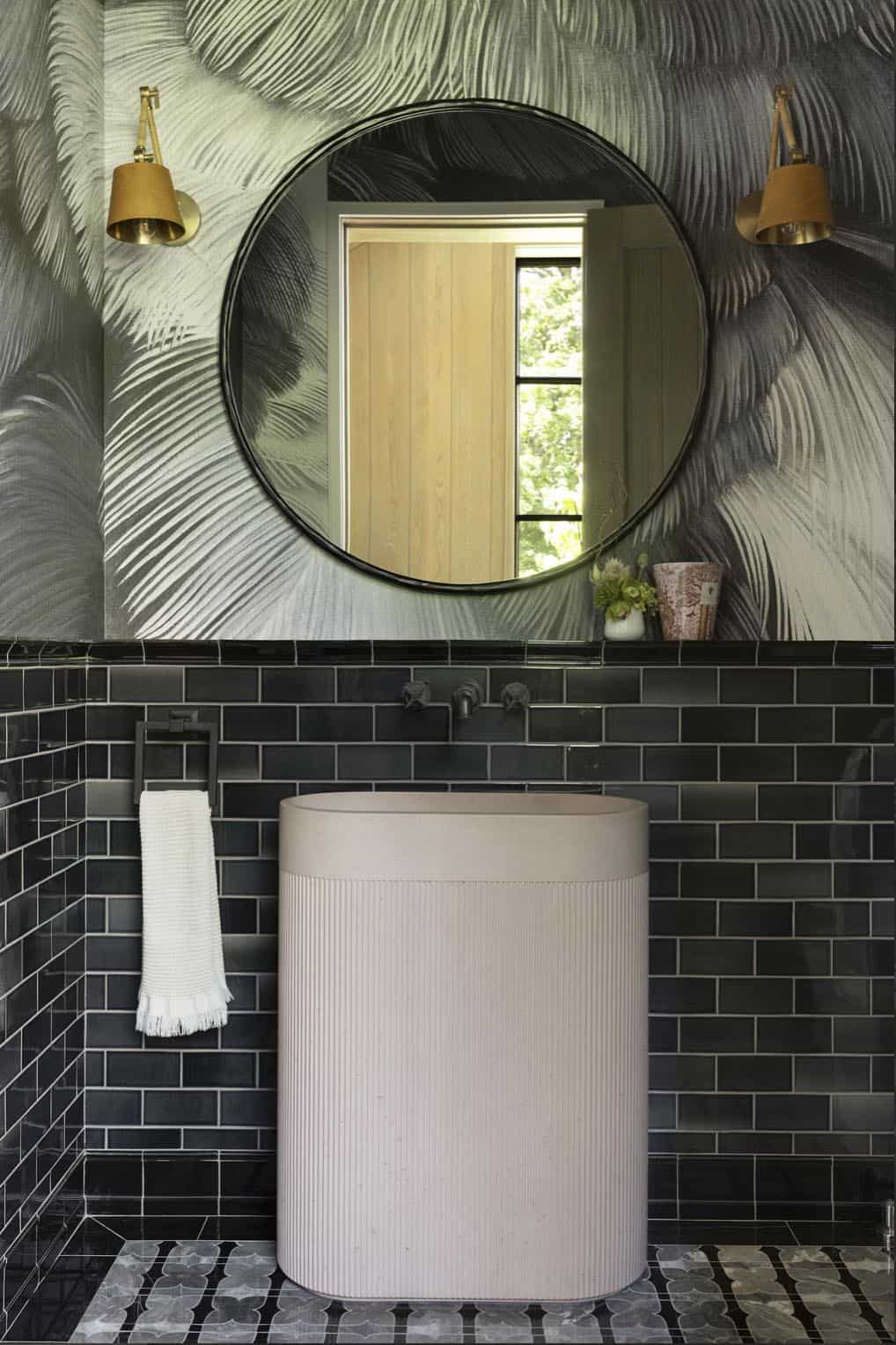 Powder room with feather-print wallpaper, round mirror, brass sconces, and dark subway tile