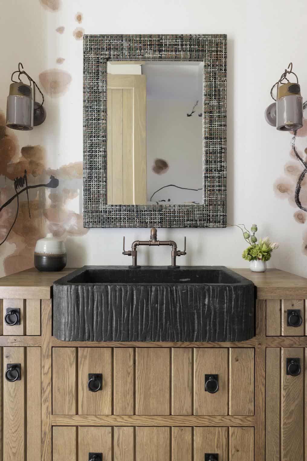 Bathroom vanity with woven-frame mirror, textured black stone sink, and paneled oak cabinetry