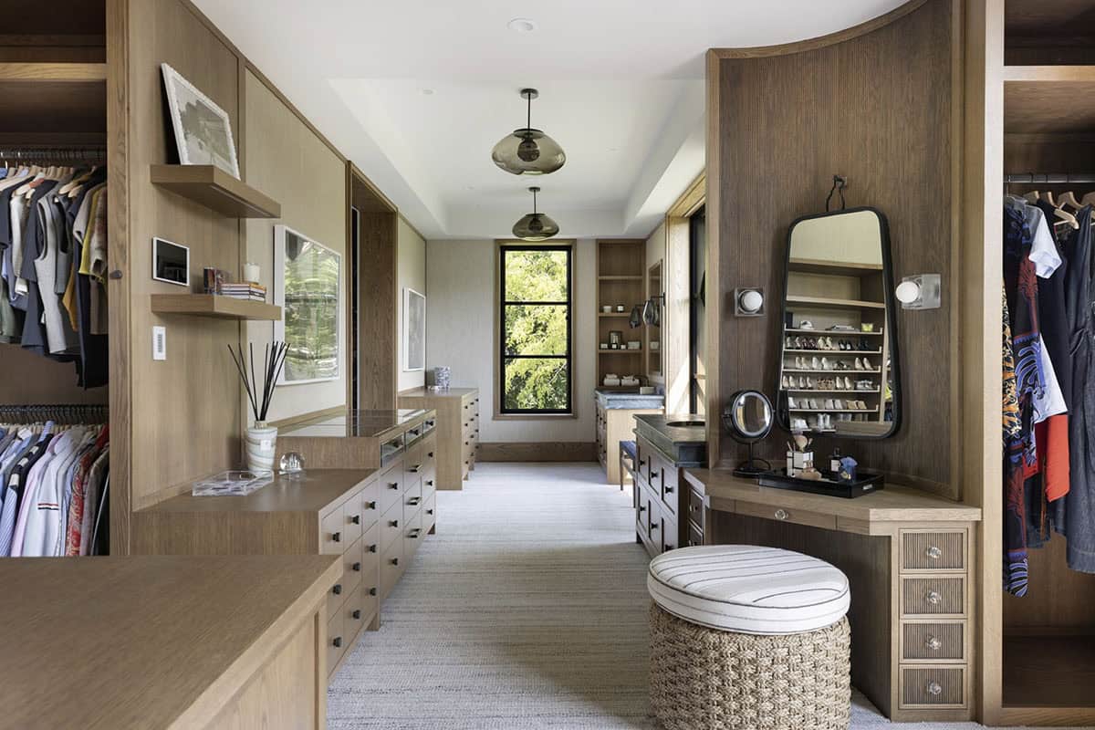 Primary closet with island, upholstered ottoman, glass pendant lights, and vanity with shoe display mirror