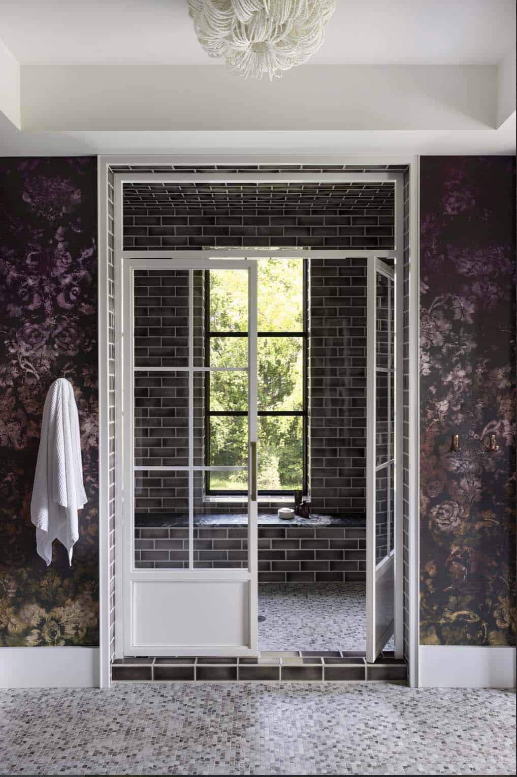 View into tiled shower with glass-paned door, window bench, and floral wallpaper surround
