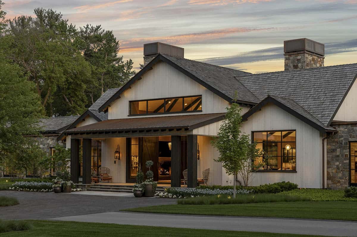 Covered entry porch at twilight with timber columns, lantern sconces, and glowing interior fireplace