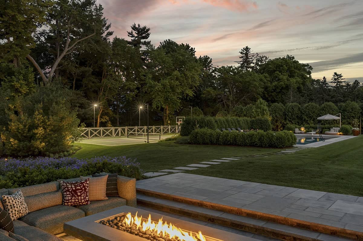 Evening view across lawn toward lit tennis court, pool, and outdoor fire pit seating