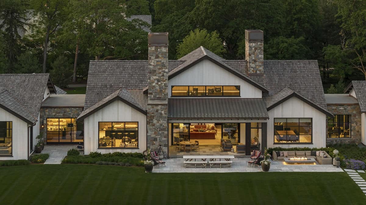 Elevated dusk view of the rear facade with stone chimneys, covered patio, and fire pit