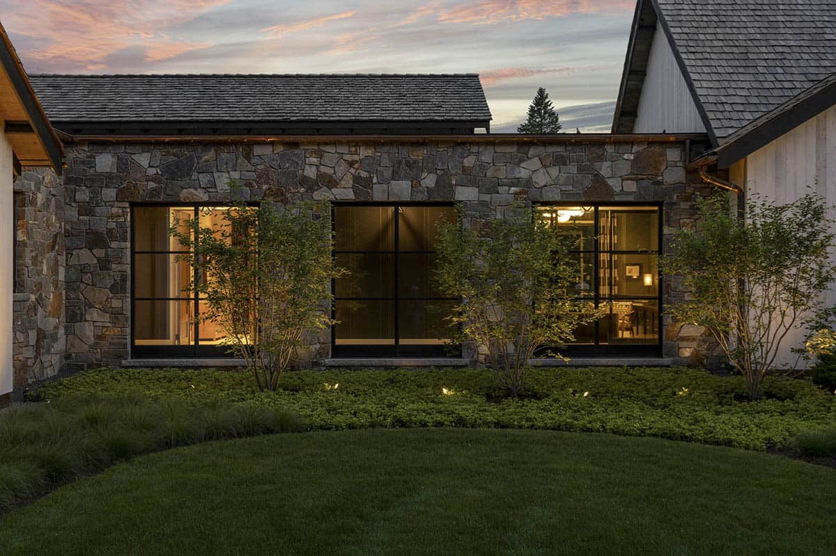 Stone-clad wing of the home lit from within, viewed across a circular lawn at dusk