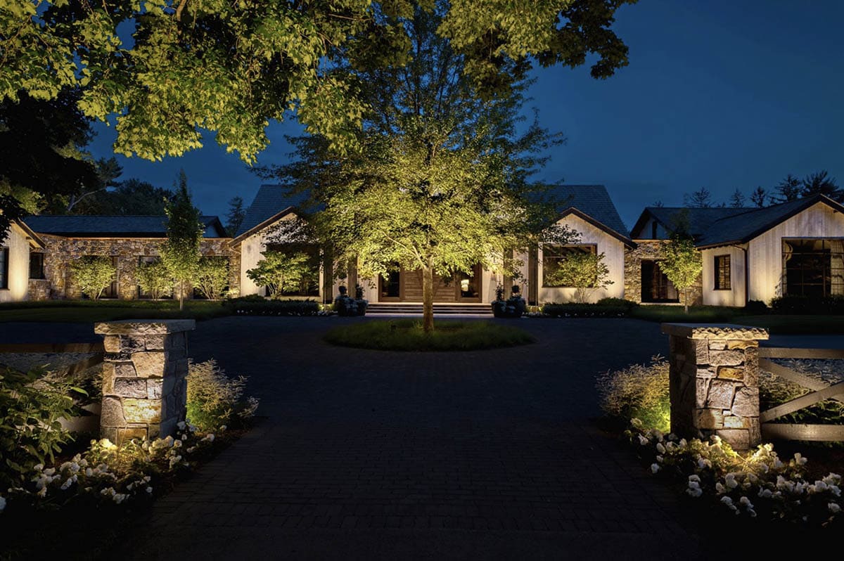 Nighttime view through stone driveway pillars toward the illuminated front entrance and center tree