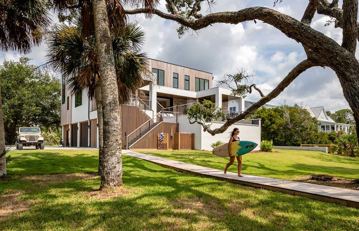 Woman carrying surfboard on boardwalk path toward modern home