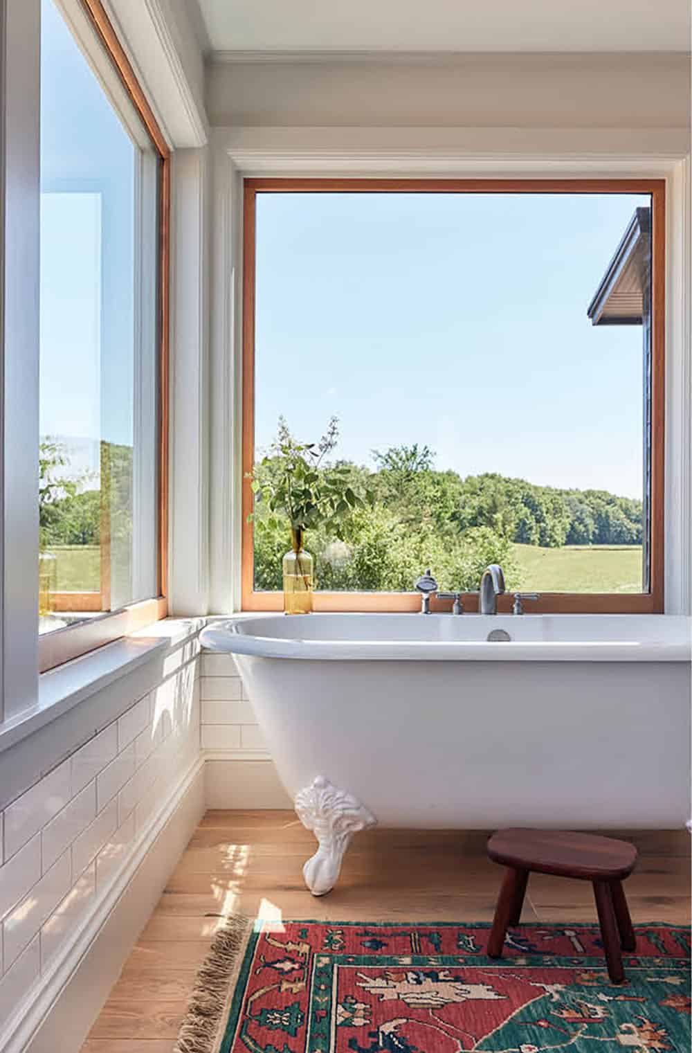 Freestanding clawfoot tub beneath a large window with wood-framed view of trees
