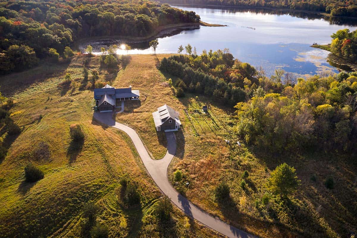 Aerial view of lake house property with driveway, outbuilding, and lake shoreline in fall
