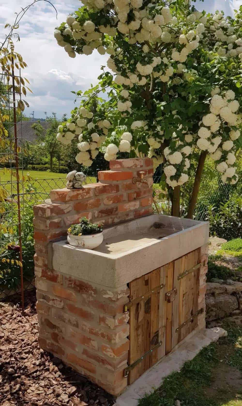 Reclaimed brick potting station with a stone sink, outdoor tap, and wood cabinet doors beneath flowering blooms