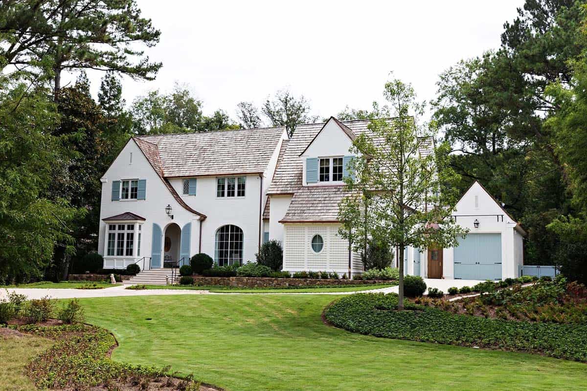 White brick Tudor-style house exterior with blue shutters, cedar shake roof, and circular driveway