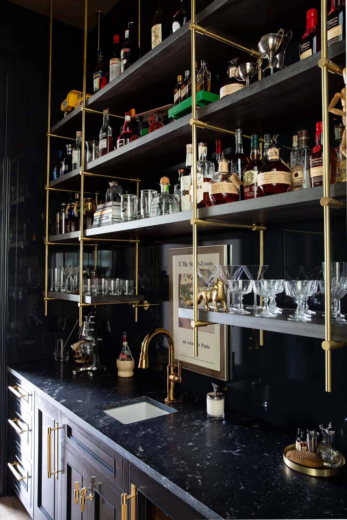 Black and brass wet bar with open shelving, liquor bottles, crystal glassware, and marble counter