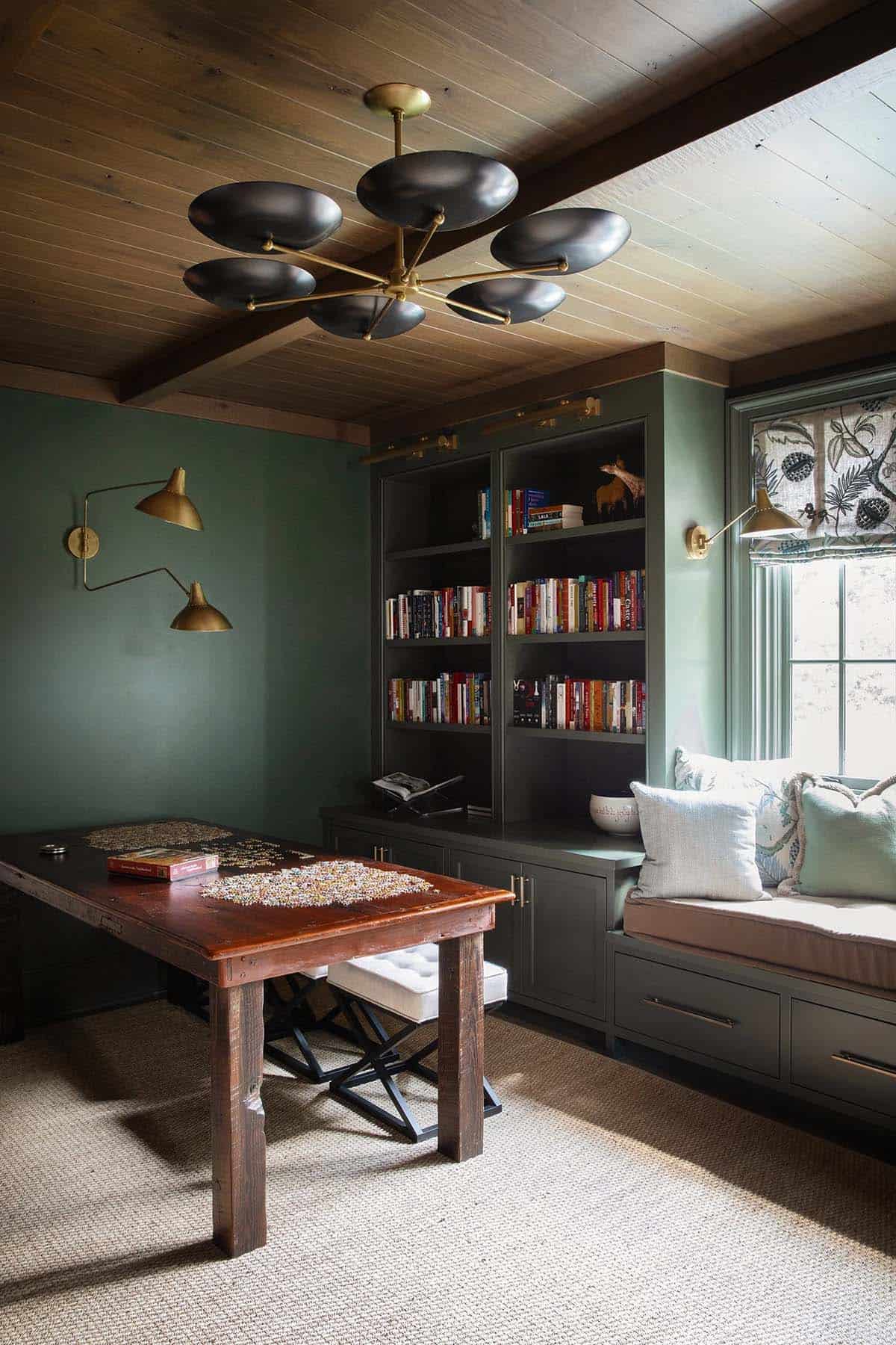 Game room with dark green walls, black and brass sputnik chandelier, built-in bookshelves, and window seat