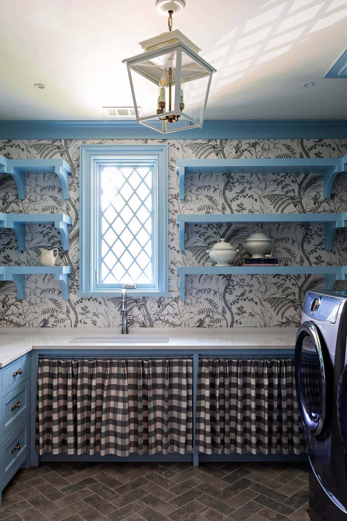 Laundry room with blue cabinetry, bird-and-botanical wallpaper, gingham skirted sink base, and brick floor