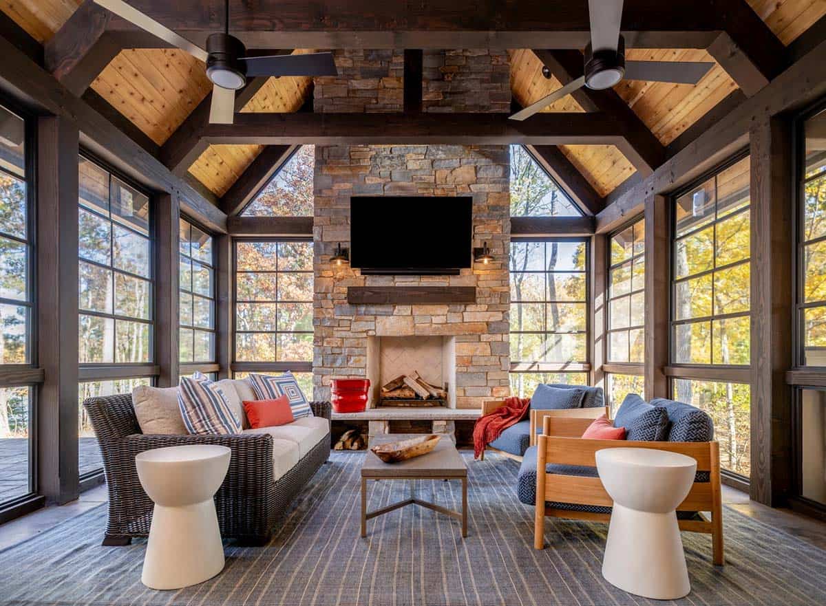 Screened porch with vaulted wood ceiling, stone fireplace, wicker sofa, and blue armchairs overlooking autumn trees