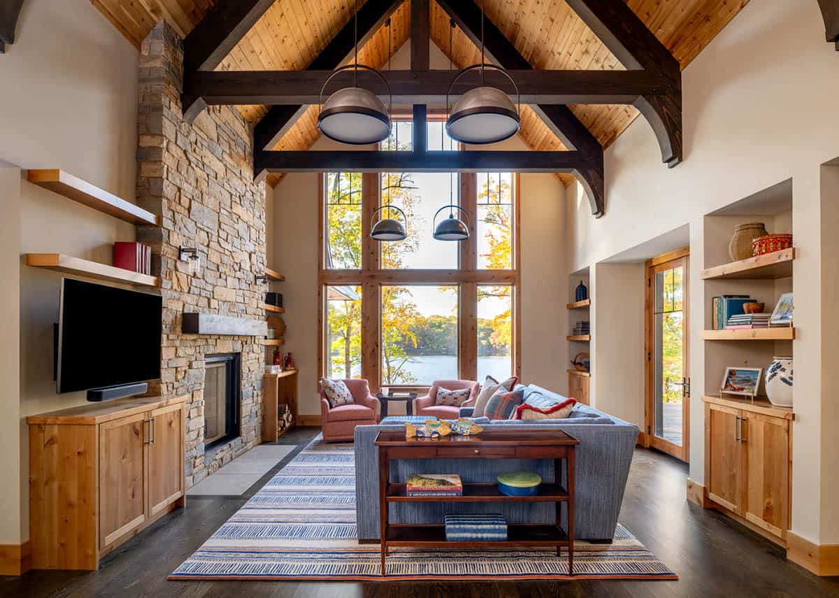 Two-story living room with stone fireplace, timber trusses, and floor-to-ceiling windows framing a lake view