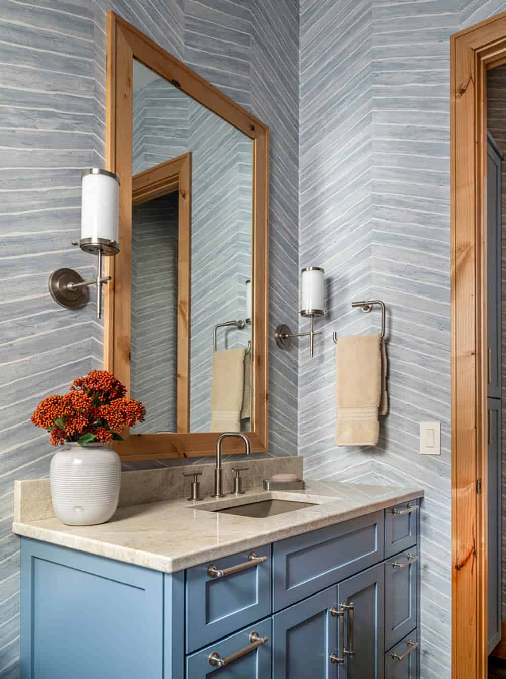 Blue bathroom vanity with wood-framed mirror, striped wallpaper, and sconce lighting