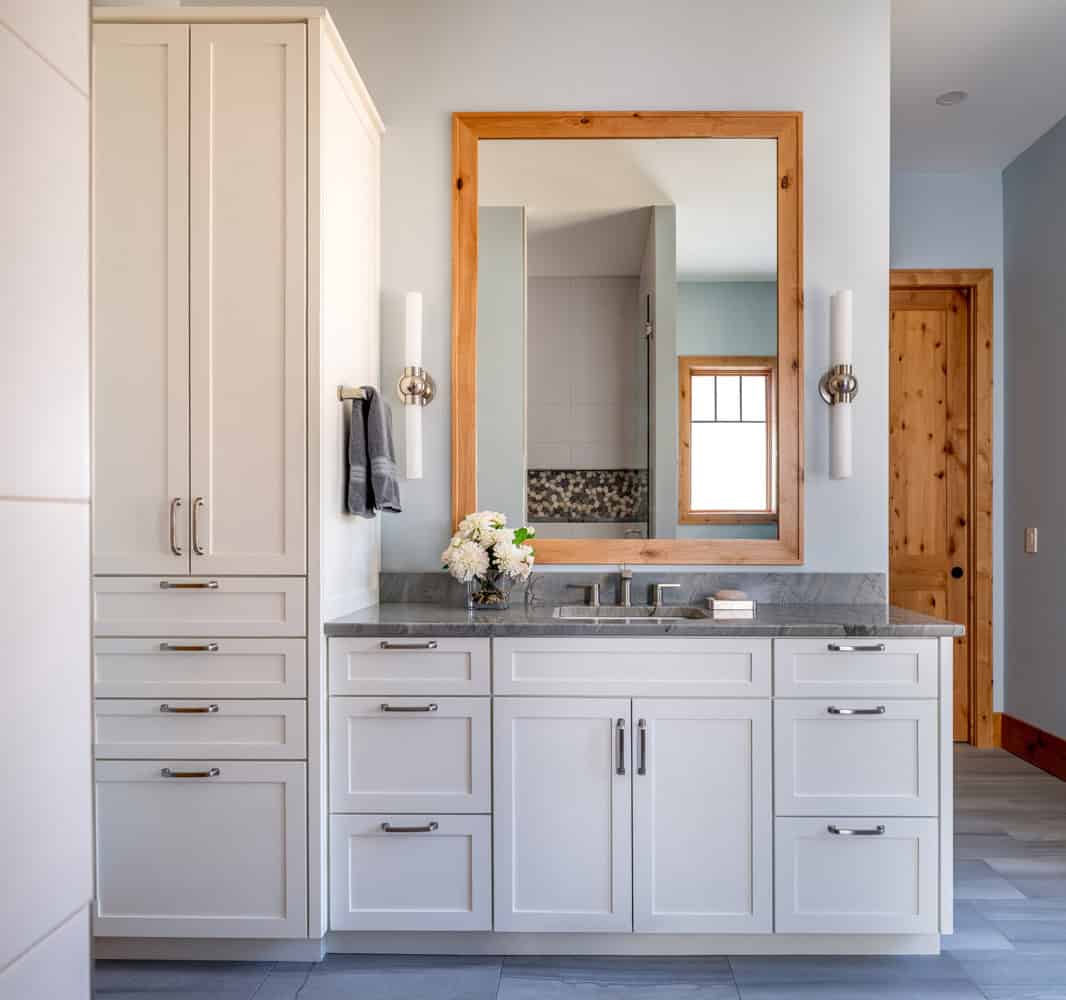 White bathroom vanity with wood-framed mirror, gray stone countertop, and linen tower