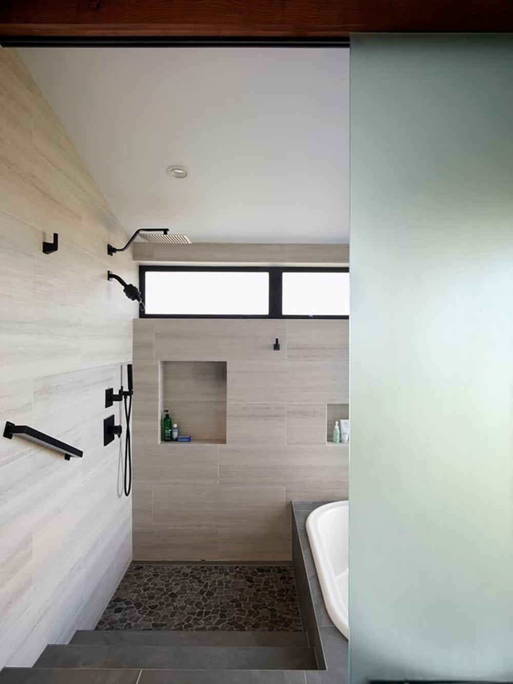 Walk-in shower with black fixtures and tiled niche beside soaking tub