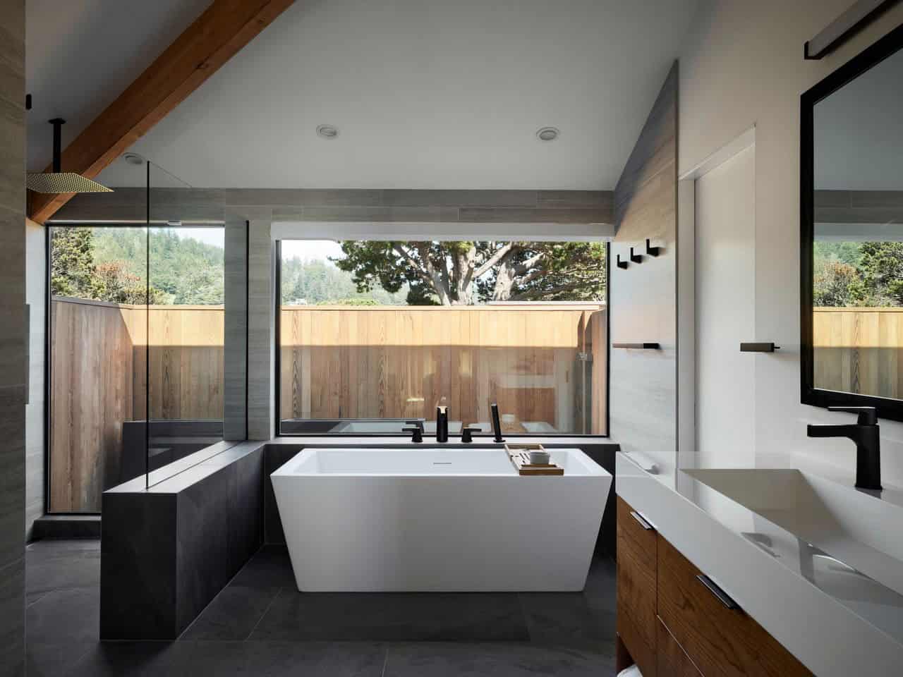 Freestanding soaking tub facing forested window view with wood fence