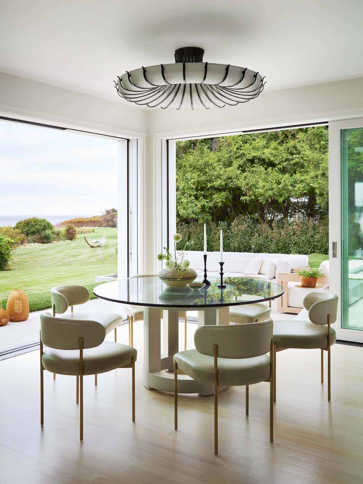 Round glass dining table with sage green chairs and sculptural pendant light opening to patio