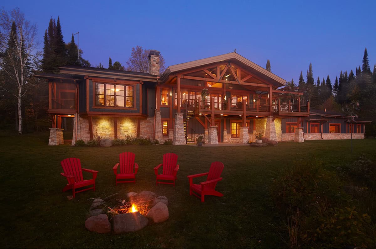 Twilight exterior view of timber-frame home with fire pit and red chairs