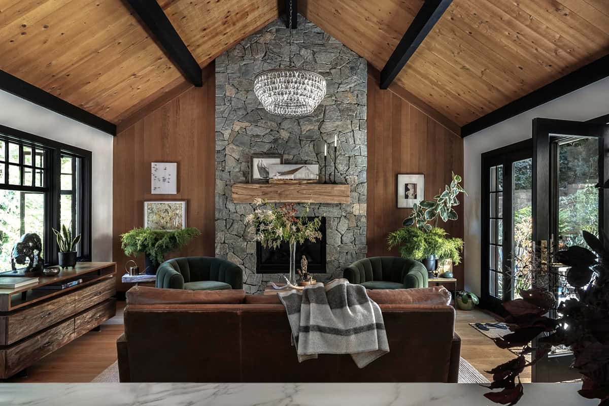 Living room with vaulted wood-plank ceiling, black beams, stone fireplace, and green velvet swivel chairs