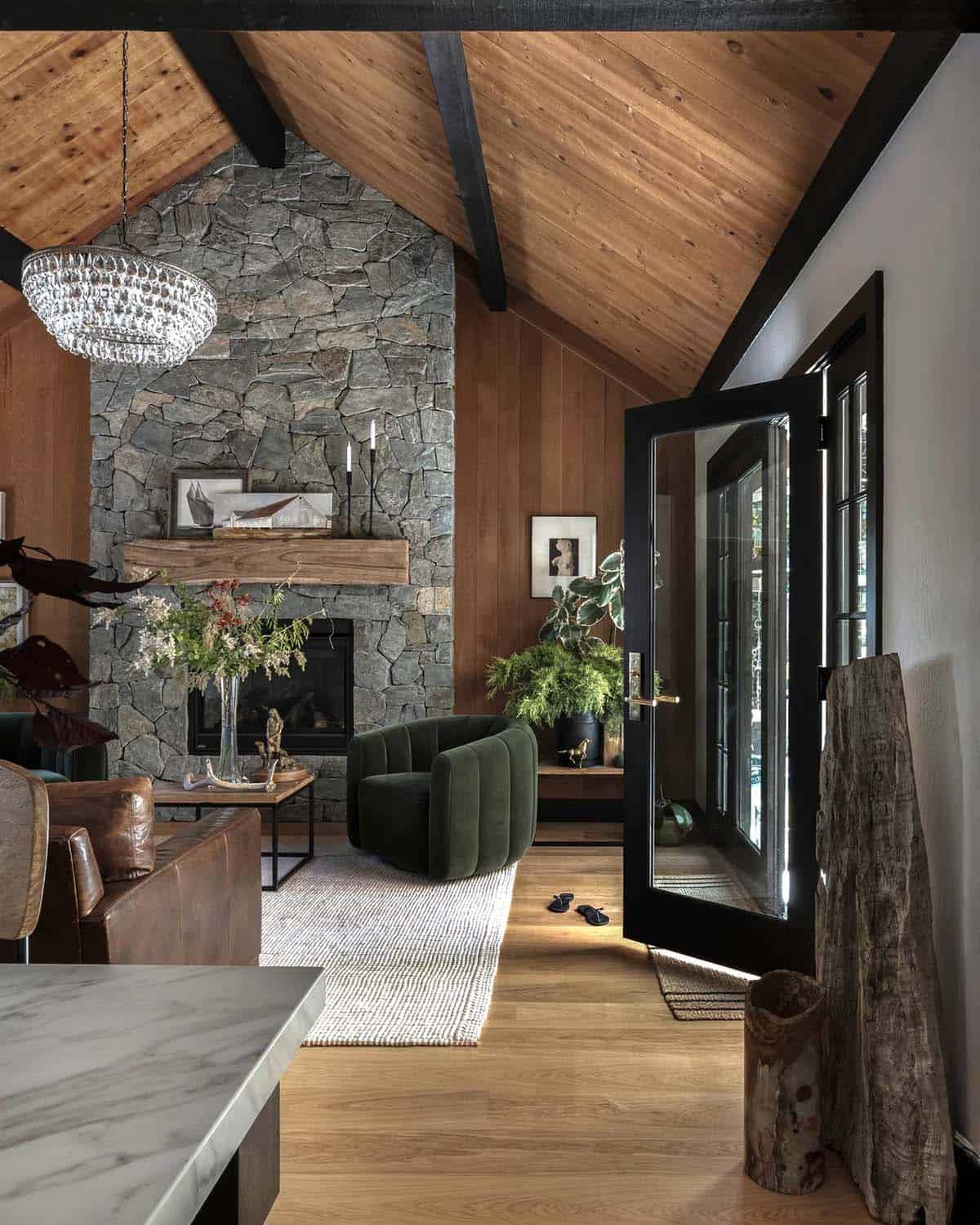Living room view toward open black-framed glass door, stone fireplace, and wood-paneled walls