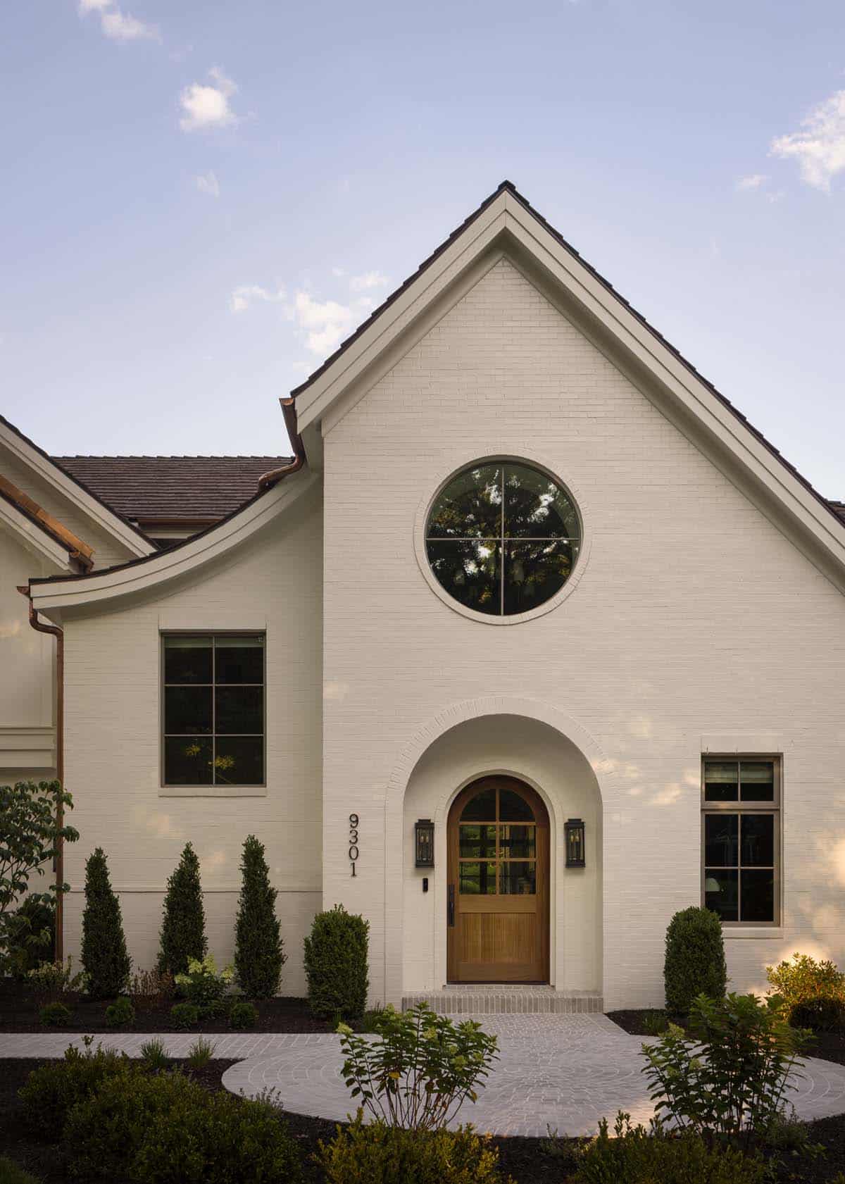Close-up of home's front gable with round window and arched wood entry door flanked by lanterns