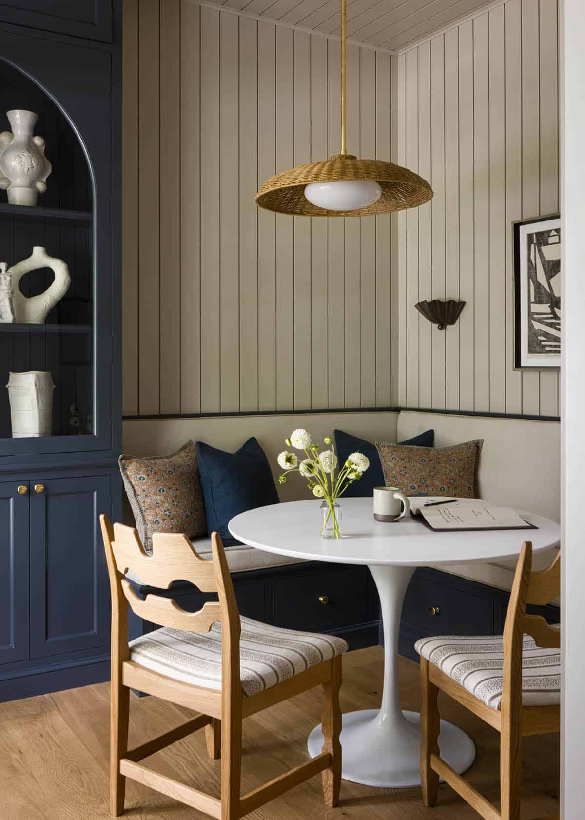 Breakfast nook with round white pedestal table, wicker pendant light, and navy built-in bench